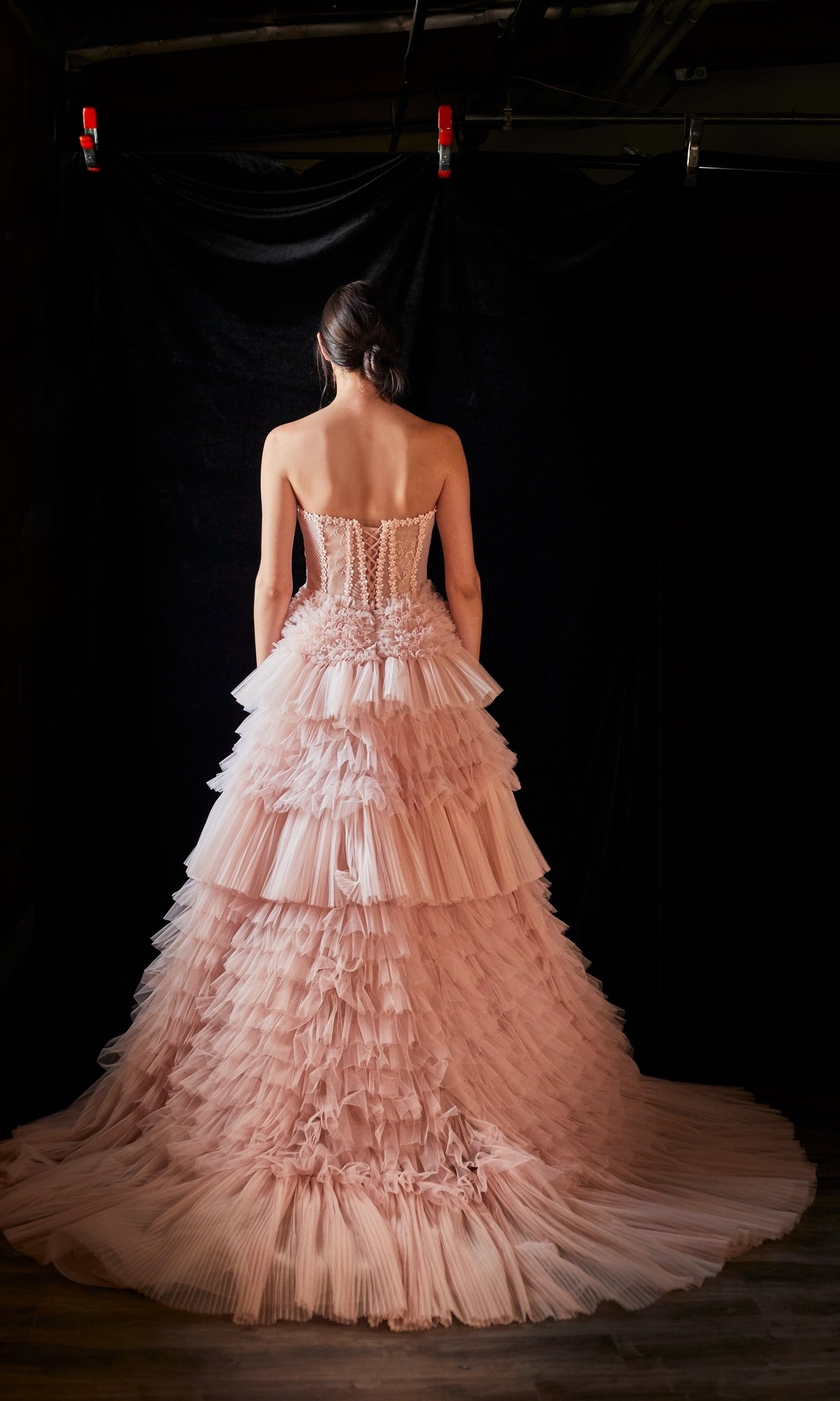 A woman faces away from the camera in the Strapless Tiered Long Prom Ball Gown A1017, its blush pink ruffled tulle tiers flowing elegantly against a dark background.